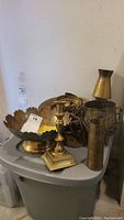 Photo showing the lot of brass items including the candle holder, bullet shell, platters, vase, and container with handles grouped together on a plastic surface against a light wall.