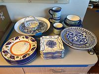 Photo showing the full assortment of ceramic dishware including plates, square plates, mugs, bowls, and the large enameled serving bowl.