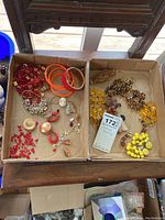 Top-down photo of two cardboard boxes filled with colorful costume jewelry including beaded necklaces, bracelets, pins, and bangles in red, orange, yellow, and brown shades.