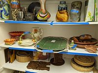 Shelves with a mix of pottery including Fitz and Floyd pitchers, baskets, vases, serving platters, bowls, and woven baskets.