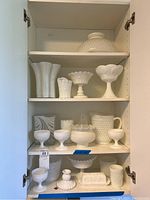 Full shelf view showing a variety of milk glass items including cups, pitcher, pedestal bowls, butter dish, and light shades arranged inside a white cabinet.