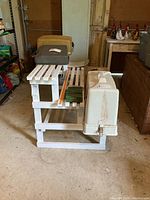 White three-tier wooden plant stand shown from the side with slatted shelves, in a garage setting with some boxes and gardening items nearby.