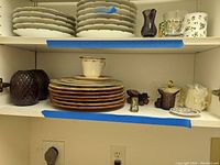 Shelf with stacked plates including several Baum Brothers Imports Tuscany Countryside plates, purple glass basket, purple glass car, abalone salt shaker, cream and sugar set, and a small cup on top of stacked plates.