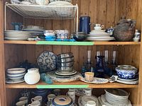 Wide view of wooden shelves displaying stacked porcelain dishes, blue and white ceramic bowls, glass vases and pitchers, and a brown ceramic jug.