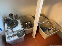 Overview of two plastic tubs containing flatware and serving pieces with candlestick holders and pewter items on top