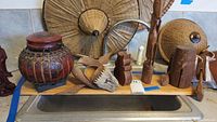 Group photo of seven wooden decorative items on kitchen counter: four carved wooden statues, one wooden carved animal mask, large round woven basket, red wooden lidded basket with woven details. One conical woven hat hangs on the wall.
