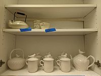 White ceramic Gracie China teapot and six covered mugs with lids featuring bird finials, alongside unmarked white teapot and four cups on shelf.