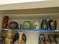 Shelf with twelve variously sized decorative vases made from art glass and ceramics. Colors range from bright swirls to earthy glazes and intricate painted details.