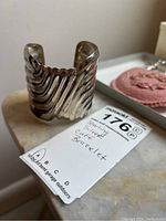 Silver cuff bracelet with ridged wave design placed on a marble surface with tag showing 'Sterling Silver Cuff Bracelet'.