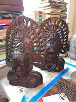 Two carved wooden tribal busts with intricate headdresses and detailed facial carvings on a table next to books and other items.