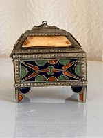 Front angled view of a small, lidded, footed metal jewelry box with multicolored blue, green, and orange enamel geometric decorations and silver metal frame.