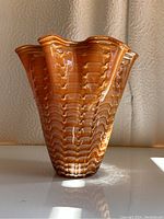 Front view of a ruffled glass vase in orange and amber hues with ribbed texture, standing on a white surface with a textured background.