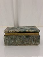 Front view of rectangular alabaster trinket box with hinged lid. Box shows natural stone pattern in greens and creams with a textured carved base and metal hinge. Lid is slightly askew and detached.