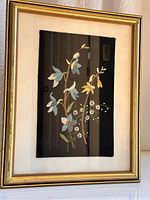 Framed piece showing detailed floral embroidery featuring blue and yellow stitched flowers on black background.