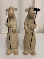 Front view of two glazed pottery sheep figurines standing upright, holding metal rods and a planting rake.
