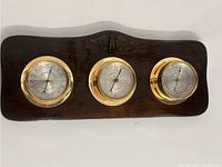 Front view of the wooden mounted weather station showing the three round instruments with brass bezels and center barometer dial.