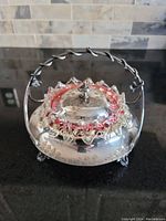 Front angle of antique silver candy dish with cranberry glass insert and floral engraved lid, showing twisted handle and butterfly accents.