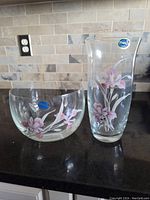 Crystal bowl and vase featuring hand-painted pink floral designs with maker's stickers visible, placed on a dark countertop with backsplash tiles in the background.