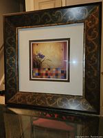 Framed print showing square artwork with flowers and checkerboard pattern inside an ornate dark frame with gold scrolling accents.