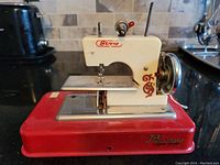 Overall view of small metal sewing machine on red base with cream upper body, showing branding, hand wheel, and sewing plate.