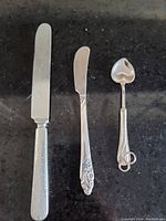 Set of two sterling silver cutting knives and one heart-shaped spoon shown from above on a black surface.