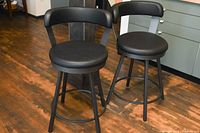Two black swivel bar stools with cushioned vinyl upholstered seats and backs, four legs with connecting footrest ring, on wooden floor.