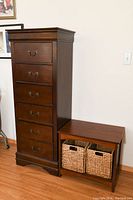 Full view of 6-drawer lingerie dresser next to bench storage unit with two wicker baskets on hardwood floor against white wall.