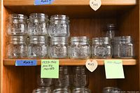 Two rows of clear Ball wide-mouth glass jars displayed on wooden shelves with sticky notes date-labeling the jars by their production period.