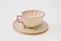 Side view showing cup and saucer, highlighting pink Neptune handle and feet, gold rim, and shell shape.