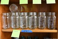 Thirteen clear wide-mouth square-sided canning jars lined up on shelf, eight Ball brand and five Kerr brand.