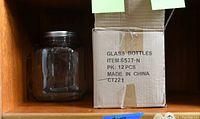 Cardboard box with one square clear glass jar with metal screw-on lid visible on the left, and the box labeled as containing 12 glass bottles visible on the right.