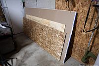 Three large construction sheets leaning against a wall: one sheet rock 3/8 inch, one plywood sheet 1/4 inch by 80x36 inches, and one strand board 1/2 inch by approximately 8x30 inches.