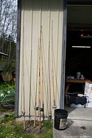 Seven fishing rods with attached reels standing against a metal panel wall outdoors in front of an open garage door.