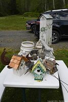 Photo of multiple bird houses and garden sculptures arranged on a white folding table outdoors, showing various shapes, sizes, and decorative themes.
