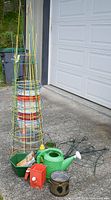 Photo showing various gardening supplies including tomato cages, peony supports, watering can, pots, and buckets arranged on driveway near garage door.