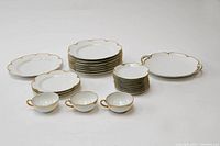 Top view of the china set arranged showing various sizes of plates, cups, saucers and two serving platters, one with handles and one without. White porcelain with scalloped edges and gold trim.