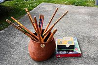 Wicker basket holding multiple wooden croquet mallets with painted colored stripes, alongside boxed bocce and cornhole games on concrete surface outdoors.