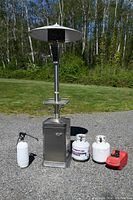 Outdoor propane patio heater with round reflector top and control panel, standing on gravel with two white propane tanks, a red portable gas tank, and a white garden sprayer positioned in front.