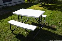 White plastic rectangular tabletop with attached benches and green metal frame, viewed from one side in outdoor grass setting.