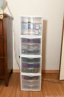 Four stacked plastic drawer units, each consisting of three drawers within a white frame, standing next to furniture against a wall.