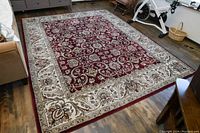 Dark red area rug on wooden floor with intricate cream border featuring floral patterns. Rug is rectangular, clean, and in very good condition.