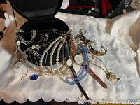 Open black jewelry box on white cloth with assorted necklaces, bracelets and watches displayed