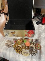 Photo showing floral decorated box open with a selection of various vintage brooches on cloth surface, displaying floral, animal, cameo and filigree designs in metal and colored stones.