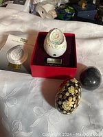 Photo of four vintage decorative eggs on fabric surface. One white porcelain egg with floral relief in red display box, black egg with gold floral design, dark stone egg, and white ceramic embossed egg. Included Cybis booklet visible.