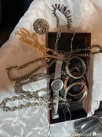 Photo showing assorted jewelry including chain necklaces, bracelets, and a wristwatch arranged inside and around a long brown jewelry box on a white embroidered cloth.