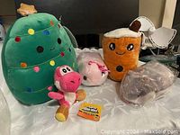 Photo of five plush toys including a green Christmas tree with colorful lights, pink Yoshi plush holding a Super Mario Bros tag, white and pink unicorn or narwhal plush, brown bubble tea plush with eyes and straw, and a wrapped grey plush toy.