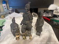 Image showing three terracotta warrior statues of varying sizes and three small brass toned peacock figurines grouped together on a cloth.