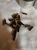 Multiple polished tube-shaped Tiger Eye gemstones with characteristic golden and brown chatoyance, laid on a white fabric surface with partial view of several small round metallic containers.