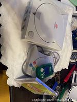 White Sega Dreamcast console with controller and green VMU resting on controller. Also shows part of game case next to controller.