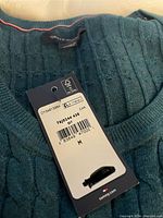 Close-up of teal Tommy Hilfiger sweater neckline showing size M tag and original price tag.